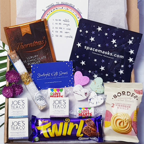 An opened gift box showing a self-heating space mask, a hot chocolate sachet, a packet of biscuits, a relaxing and nourishing aromatherapy bath salts/facial steamer, a set of miniature heart bath bombs, two small packets of love hearts, a Cadbury Twirl, a 5 things to remember today positivity card, and a trio of tea