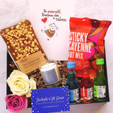 An opened gift box showing three bottles of fruit Vodka shots, a handmade Belgian chocolate slab, a scented candle, a packet of sticky cayenne nut mix and a positivity card