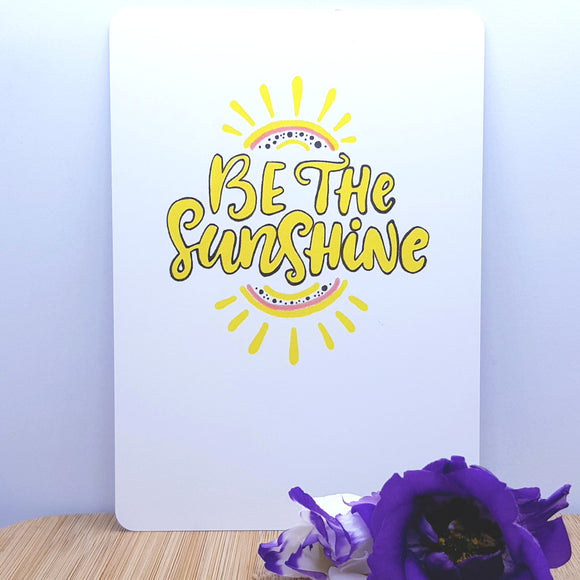 An A5 card showing a sun with be the sunshine written on it, decorated with a purple flower in front