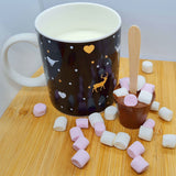 A mug of hot milk next to a hot chocolate stirrer, scattered with mini marshmallows