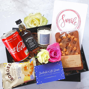 An opened gift box showing a can of coke, a bottle of Jack Daniels, a slab of Belgian chocolate, a scented candle, a smile positivity print and a packet of biscuits