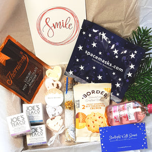 An opened gift box showing a bottle of Gordon's pink gin, a self-heating space mask, a hot chocolate sachet, a packet of biscuits, a soothing aromatherapy bath salts/facial steamer, a set of miniature heart bath bombs, a smile positivity card, and a trio of tea