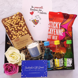 An opened gift box showing three bottles of fruit Vodka shots, a handmade Belgian chocolate slab, a scented candle, a packet of sticky cayenne nut mix and a positivity card